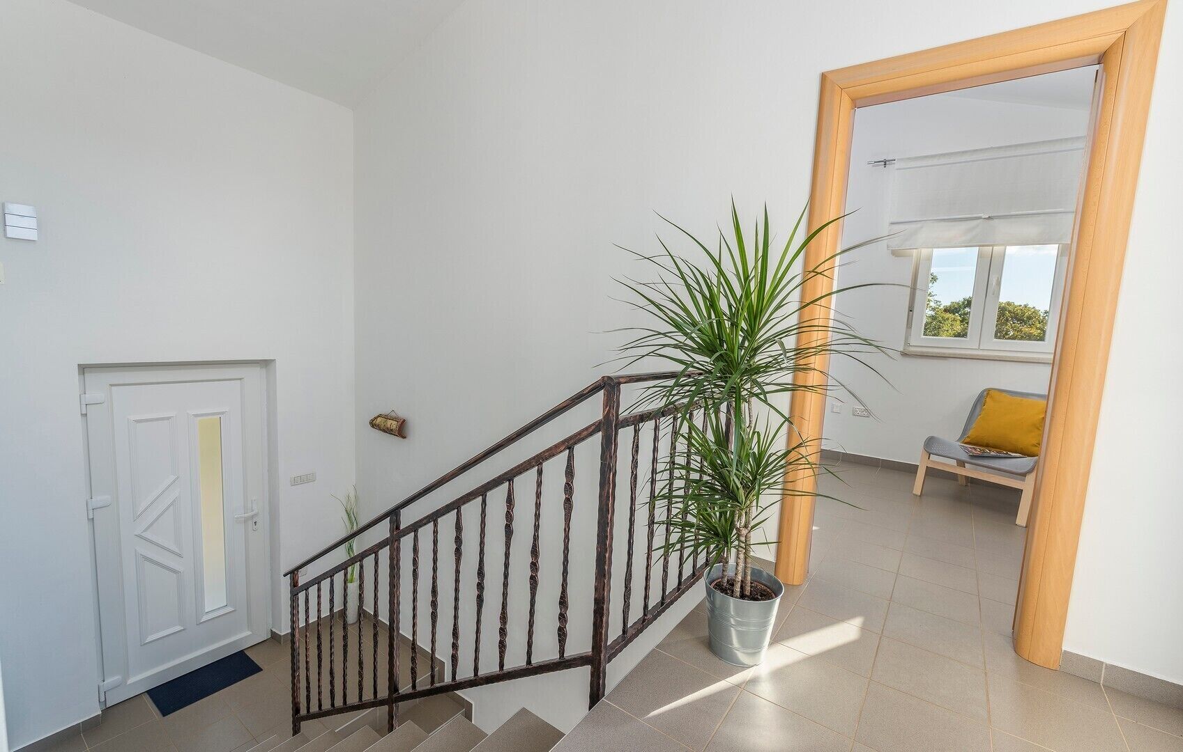 Una escalera con barandilla metálica, una planta en maceta, una puerta blanca y vista de una habitación con ventana y una silla.