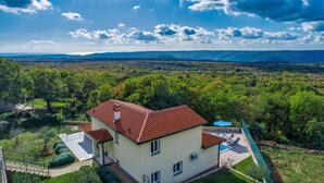 Ein zweigeschossiges Haus mit rotem Ziegeldach, einem Swimmingpool und Blick auf eine bewaldete Landschaft.