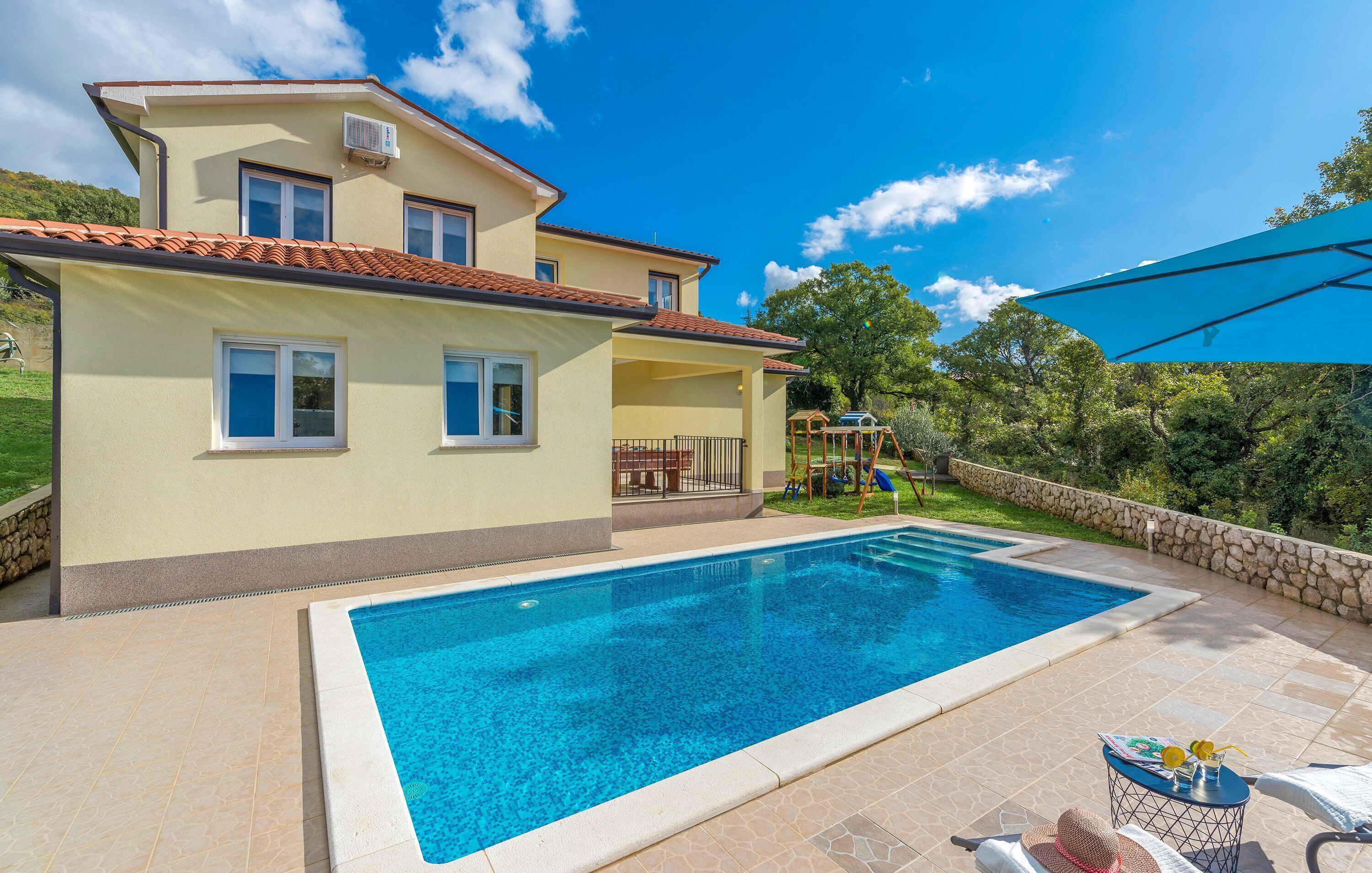 A two-story house with a swimming pool, outdoor furniture, and a stone wall.