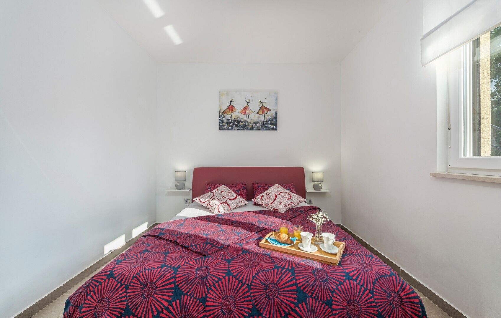 A small, neatly arranged bedroom with a bed, bedside lamps, a window, and a breakfast tray on the bed.