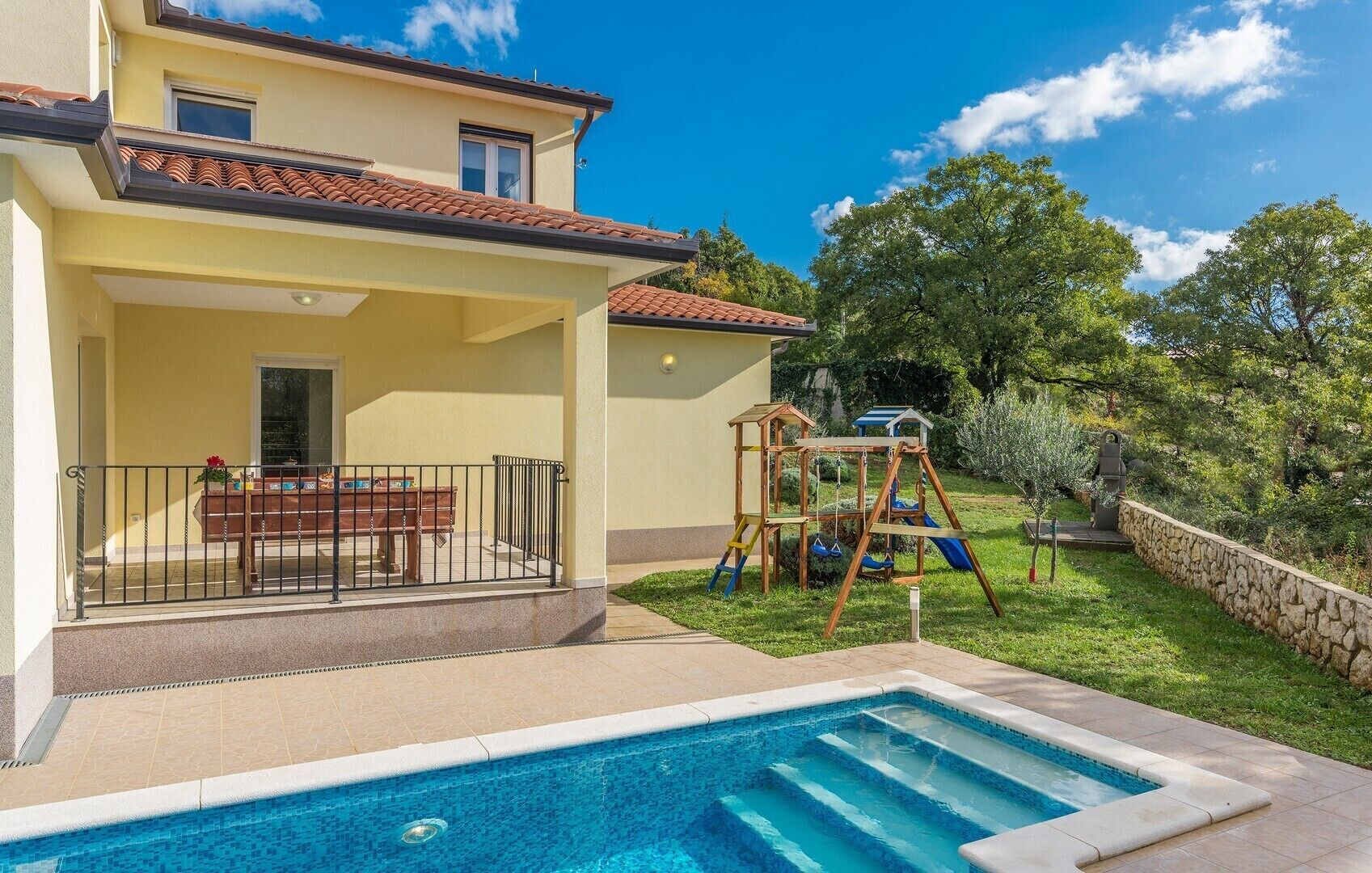 A two-story house with a swimming pool, a patio with a table and chairs, and a children's play structure.