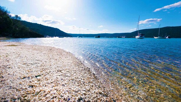 Ein Kiesstrand mit klarem Wasser und Booten in der Ferne.