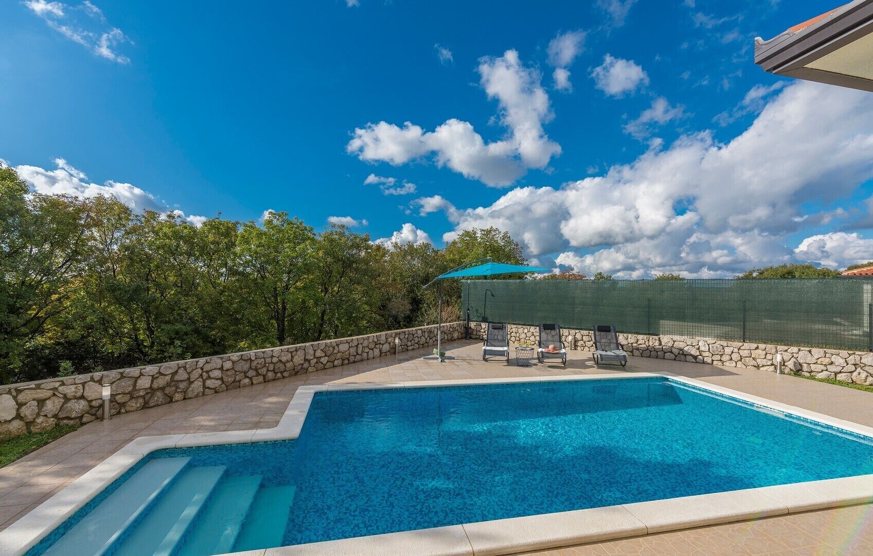 A swimming pool with a stone wall and a covered area with lounge chairs.