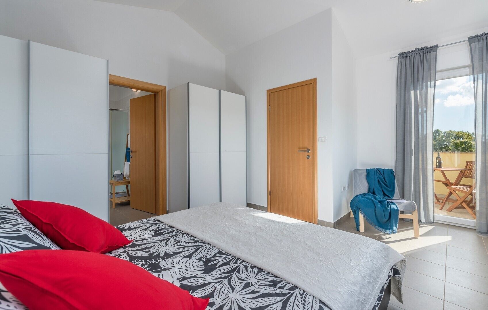 A bedroom with a bed, a wooden wardrobe, a door, and a balcony with a table and chairs.