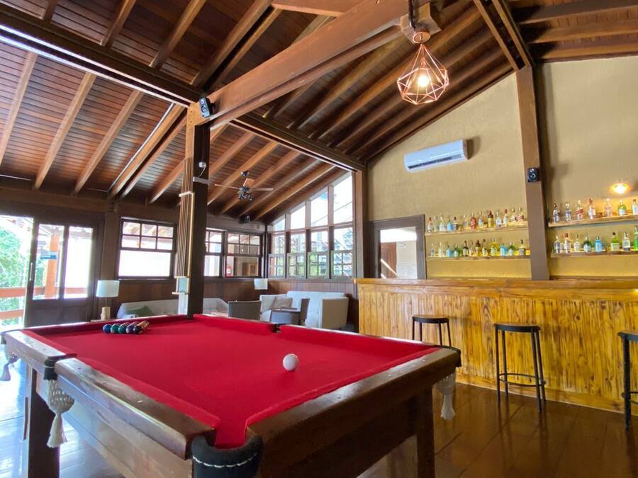 Una mesa de billar con superficie de fieltro rojo y una bola blanca, una zona de bar con diversas botellas, y un techo de madera con vigas a la vista.