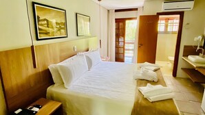Quarto de hotel com uma cama grande, cabeceira de madeira, mesa de cabeceira com telefone, banheiro com pia e espelho, e porta que dá para uma área externa.