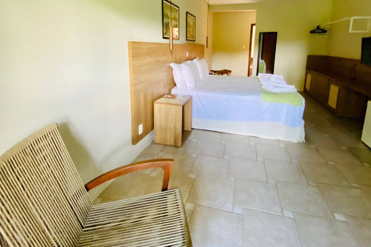 A hotel room with a bed, a chair, a bedside table, an air conditioning unit, and a door leading to another room.