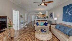 A spacious living room with a wooden floor, a ceiling fan, a sofa with blue cushions, a coffee table, and a television.