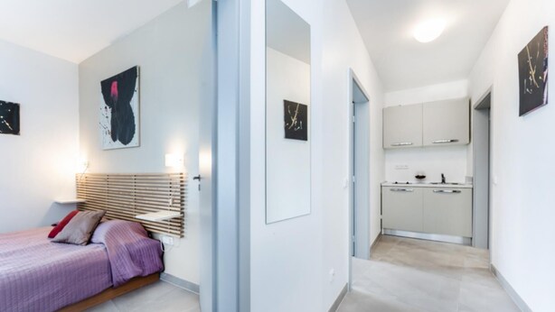 Une chambre moderne avec un lit, une table de chevet et une lampe fixée au mur. Un couloir mène à une cuisine équipée de placards et d’un plan de travail.
