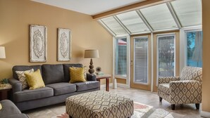 Un salon avec un canapé gris, un pouf à motifs et un fauteuil, une lampe, ainsi que deux cadres décoratifs fixés au mur.
