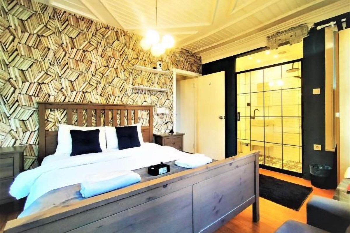 A bedroom with a wooden headboard, a bed with white bedding, a bedside table, and a large window with a view.