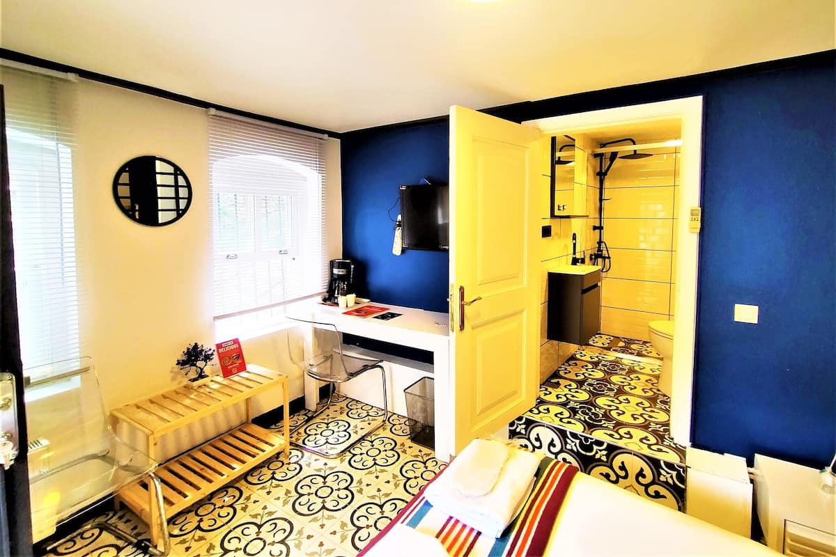 Una habitación con cama, escritorio, silla, radiador, ventana con persianas y baño con ducha e inodoro.