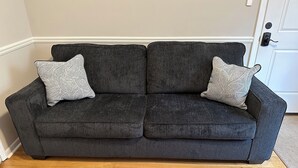 Un canapé gris foncé agrémenté de deux coussins décoratifs à motifs, dans une pièce au plancher en bois et dont le fond est marqué par une porte blanche.