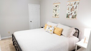 Un lit bien fait, recouvert d’une couette blanche, avec deux oreillers à motifs floraux et une petite table de chevet sur laquelle se trouve une lampe.