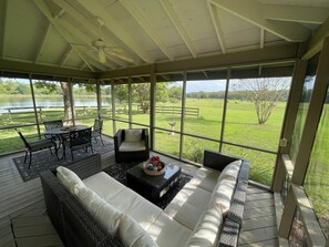 A covered outdoor seating area with wicker furniture, a table with a centerpiece, and a view of a grassy field and lake.