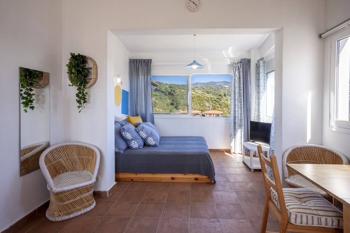 Un dormitorio con una cama, dos sillones de mimbre, una mesa de madera, un televisor y un ventanal con vistas a las colinas.