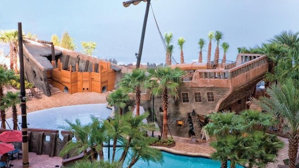 A pool area with a ship-themed structure, palm trees, and a clear sky.
