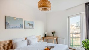A hotel room with a bed, a wooden nightstand, a small table with a vase of flowers, and a large window with a view of buildings. - Brussels Royal Design Residence (Brussels)
