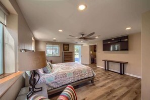 A bedroom with a bed, a ceiling fan, a TV mounted on the wall, and a window.