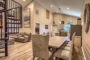 A spacious living area with a dining table, cushioned chairs, a sofa, a staircase, and a kitchen in the background.
