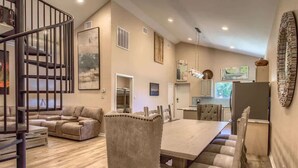 A spacious living area with a dining table, cushioned chairs, a sofa, a staircase, and a kitchen in the background.