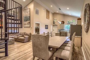 A spacious living area with a dining table, cushioned chairs, a sofa, a staircase, and a kitchen in the background.