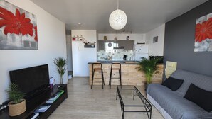 Un salon moderne avec un canapé, une table basse et un téléviseur. On aperçoit une cuisine au fond, avec un coin bar.
