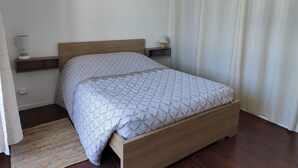 Une chambre à coucher avec un lit en bois, une table de chevet avec une lampe, une œuvre d’art accrochée au mur et une fenêtre avec des rideaux.