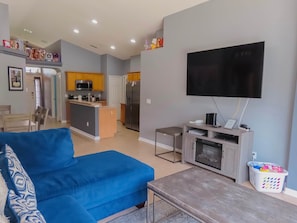A living room with a blue sofa, a TV, a small table, and a kitchen in the background.