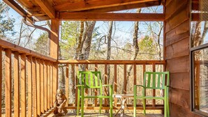 A wooden deck with two green chairs and a table, surrounded by a wooden railing, overlooking a forested area.