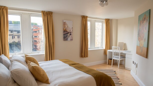 Un dormitorio con una cama grande, un escritorio y una silla. Hay ventanas con cortinas, un cuadro en la pared y vistas a edificios.