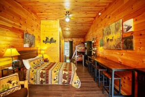 A log cabin-style room with a bed, a desk, and chairs, featuring wooden walls and ceiling, and artwork depicting wildlife. - Bigfoot Lodge Room Three - Outdoor Adventures (Benton)