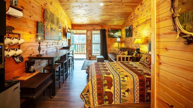 Ein gemütliches Blockhaus-Interieur mit einem Holzesstisch, Bänken, einer Couch und einem Bett mit einem gemusterten Überwurf.