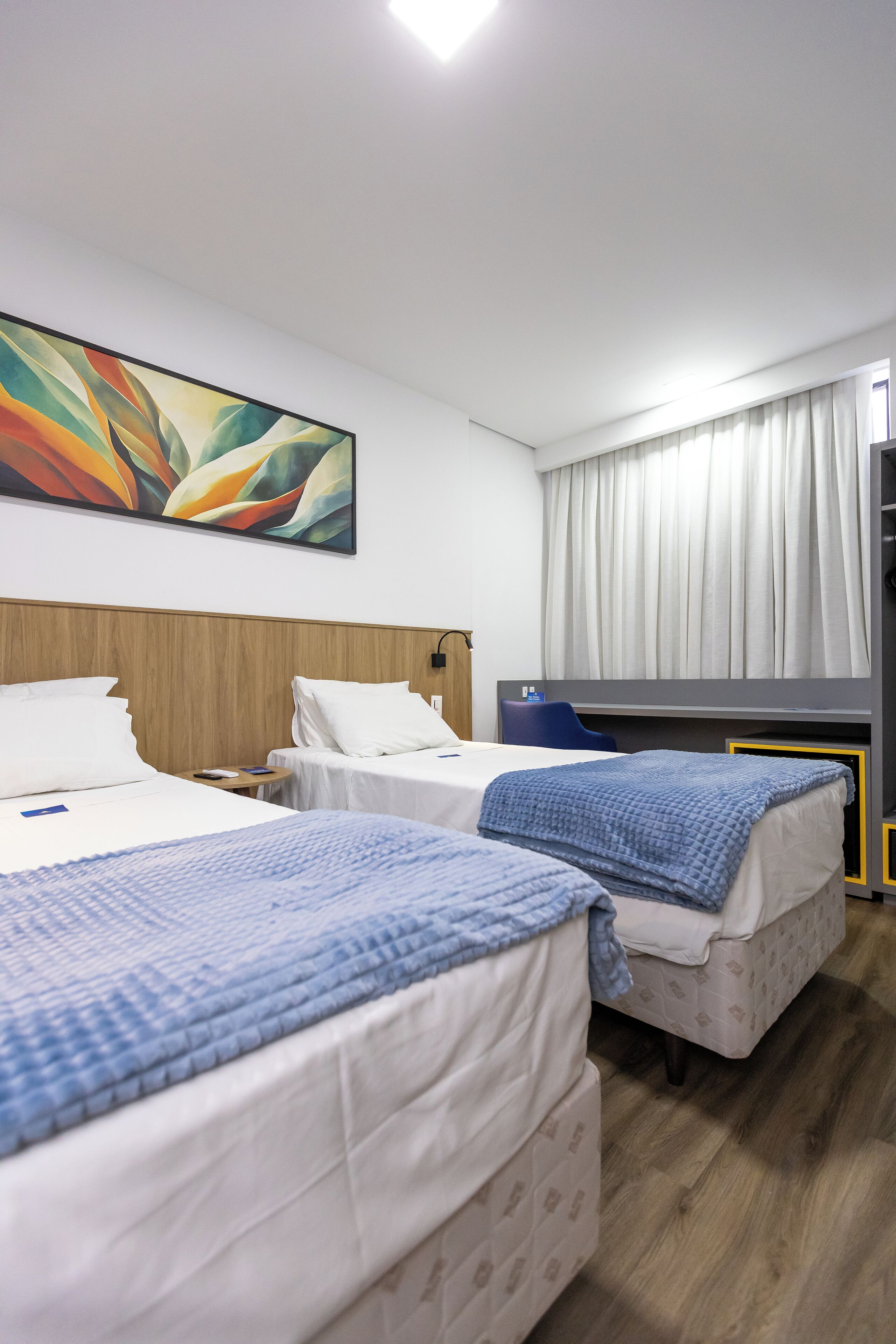 Quarto de hotel com duas camas, cabeceira de madeira, um quadro na parede e uma janela com cortinas.