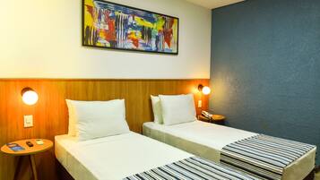 Habitación de hotel con dos camas, cabecero de madera, cuadro abstracto de colores, una mesa redonda con un teléfono y un libro, y una mesita de noche con una lámpara.