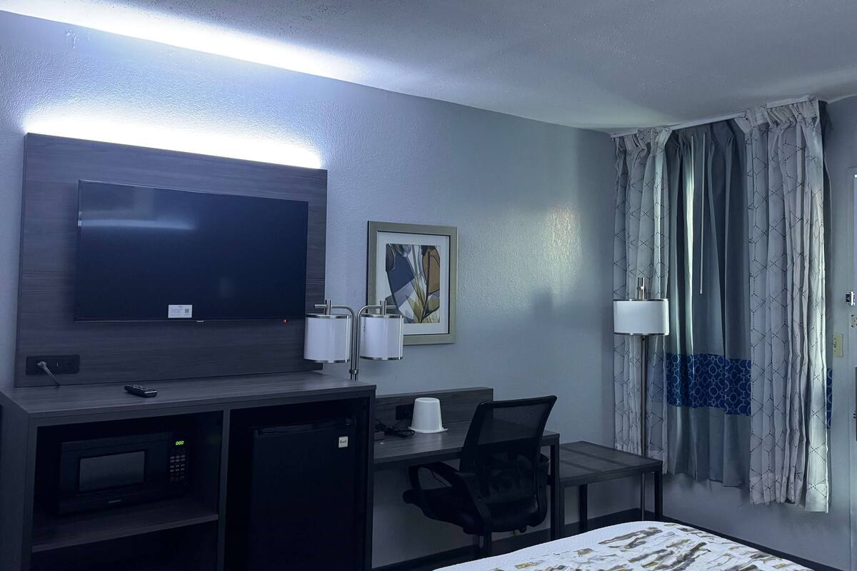 Hotellihuone, jossa on taulutelevisio, työpöytä tuolilla, mikroaaltouuni, pieni jääkaappi, yöpöytä ja sänky.