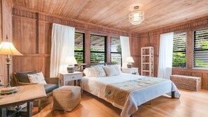 A wooden-paneled bedroom with a bed, two bedside tables with lamps, a chair, a small table, and a bookshelf. - Five Bed, Five Bath Tropical Paradise With A/C, Hot Tub, & Beach Walk (Koloa)