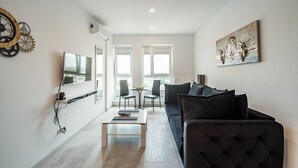 A modern living room with a black sofa, a coffee table, and a TV mounted on the wall. There are two windows with blinds, a painting on the wall, and a clock with gears. - Cozy Penthousestudio Cityview W12 (Bucharest)