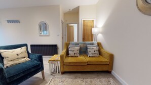 Ein Wohnzimmer mit einem blauen Sofa, einem senfgelben Sofa und einem kleinen Tisch. Ein Heizkörper, eine Lampe und ein Fenster mit einem Spiegel darüber sind ebenfalls zu sehen.
