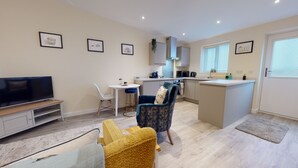Eine moderne Wohnung mit Wohnzimmer, Essbereich und Küche. Es gibt einen Flachbildfernseher, ein Sofa, einen Sessel und einen kleinen Tisch. Die Küche ist mit einem Spülbecken, einem Herd und einer Mikrowelle ausgestattet.