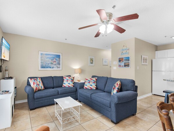 Un salon avec un canapé bleu, une table basse blanche, un ventilateur de plafond et un réfrigérateur blanc.