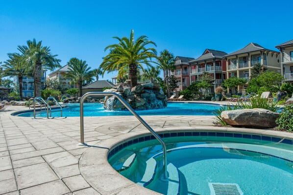 A swimming pool with a stone fountain, palm trees, and a row of multi-story buildings with balconies.
