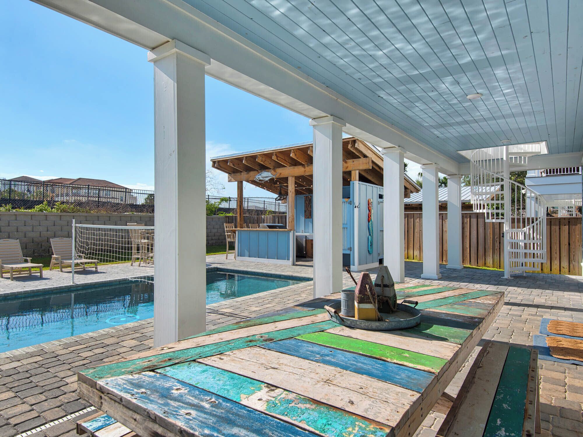 A covered outdoor swimming pool area with a wooden deck, a barbecue station, and a spiral staircase.