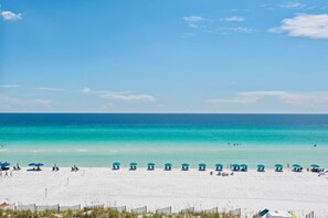 A beach with white sand and clear turquoise water. There are several blue umbrellas and lounge chairs along the shoreline. - Gulf Front, Gated Community (Destin)