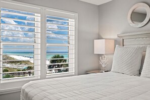 A bedroom with a large window, a bed with white bedding, a bedside lamp, and a round mirror on the wall. - Gulf Front, Gated Community (Destin)