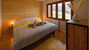 A bedroom with a wooden headboard, a bed with patterned bedding, a bedside table with a vase, and a window with a view of the outdoors. - Cosy*SPA*Modern- at Ô JurassiK (Coteaux du Lizon)