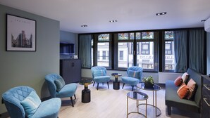 Un salon moderne avec des fauteuils bleus, un canapé et une table basse. Un tableau représentant un paysage urbain encadré est accroché au mur, ainsi qu’un meuble et une fenêtre avec des rideaux.
