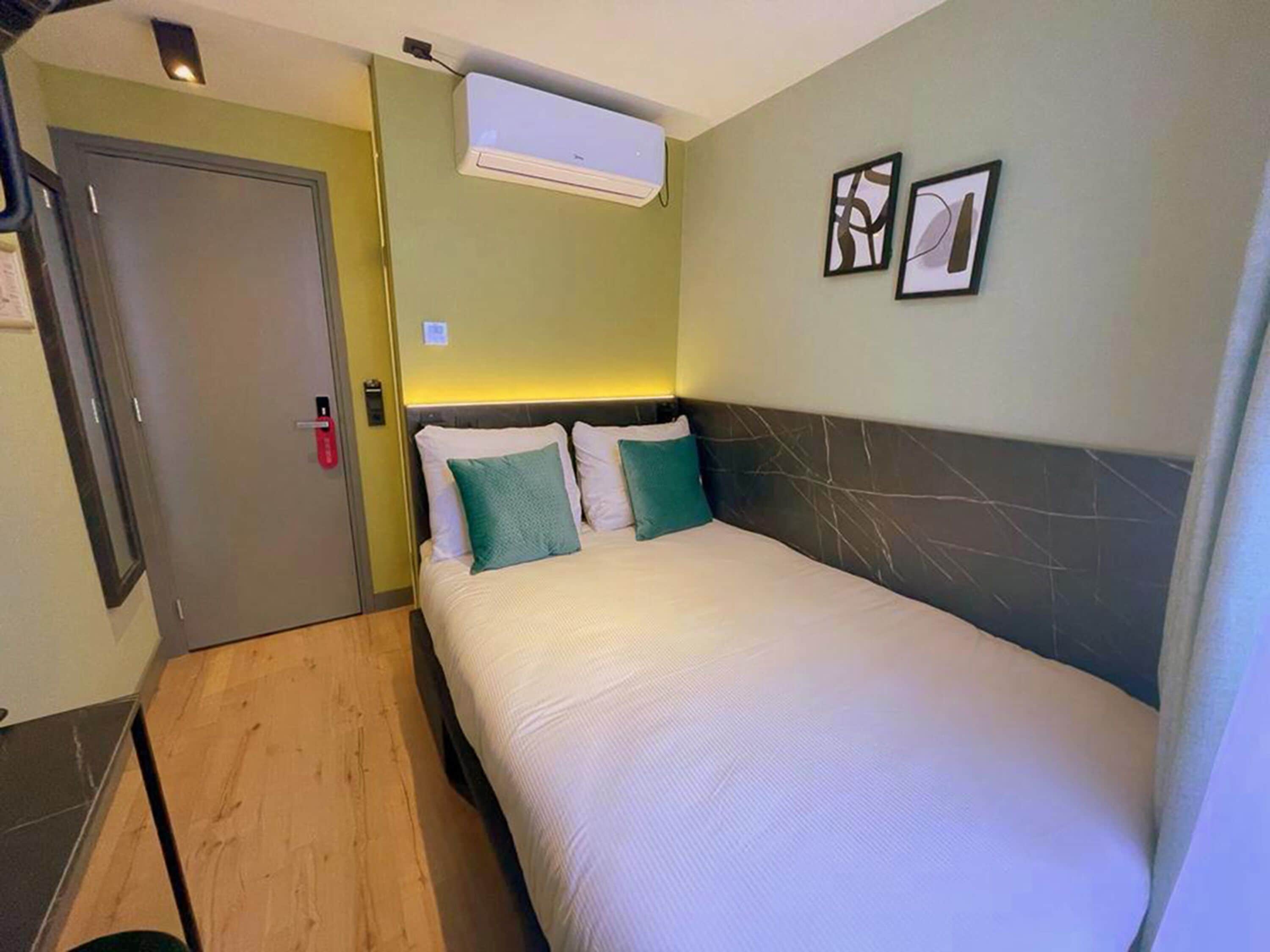 A hotel room with a bed, a door, an air conditioning unit, and two framed pictures on the wall.