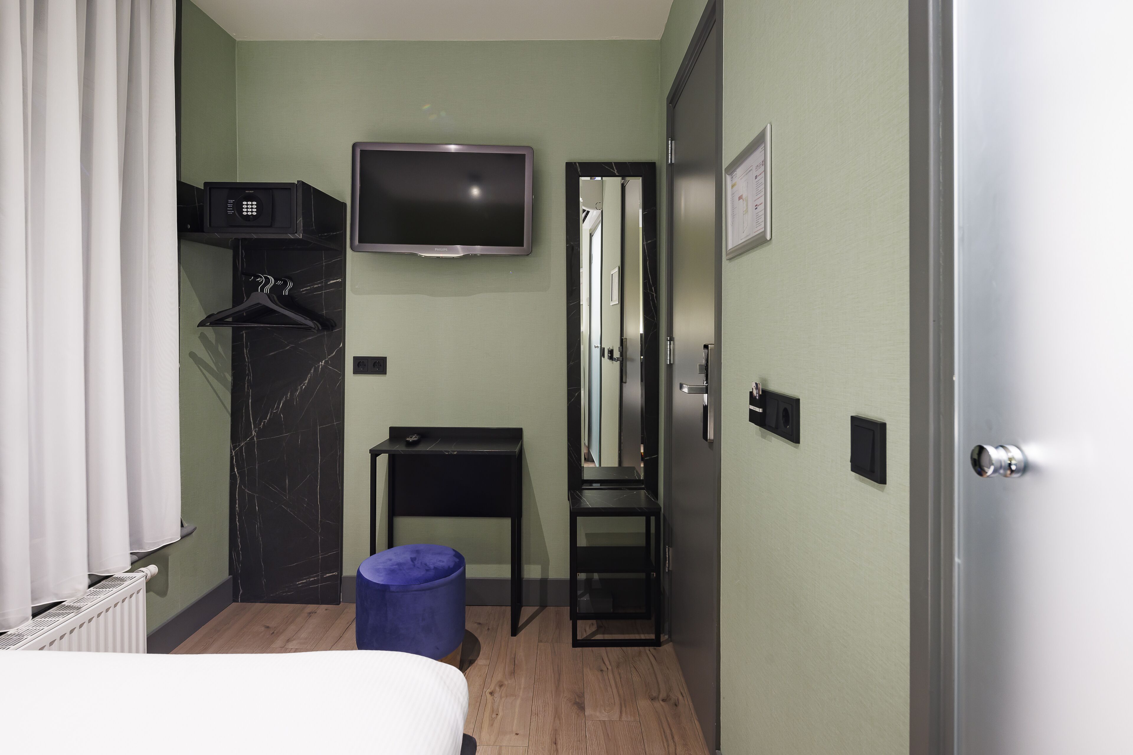 A compact hotel room with a bed, a TV mounted on the wall, a small table, a mirror, and a wardrobe.