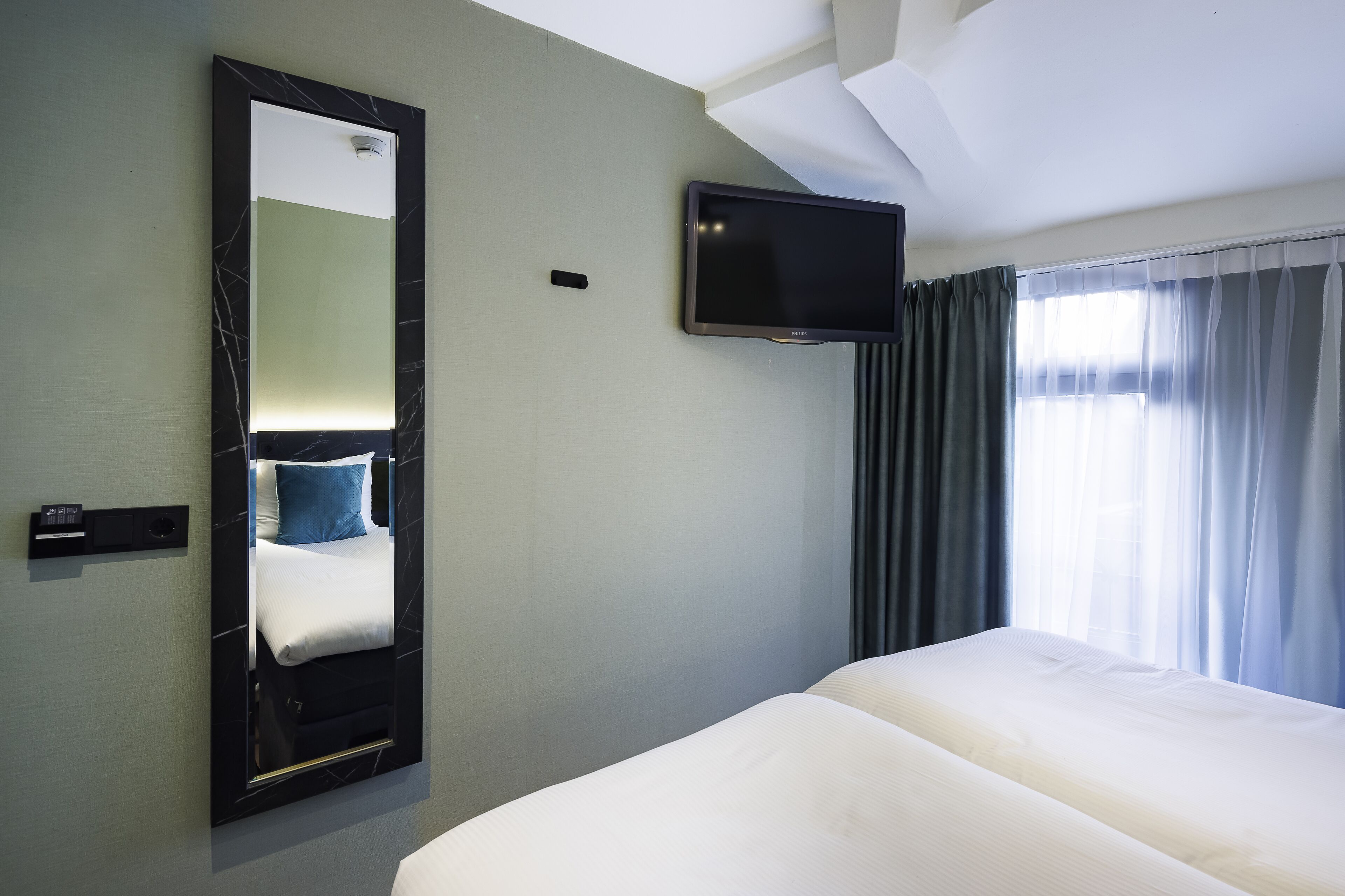A hotel room with a bed, a large mirror, a flat-screen TV, and a window with curtains.